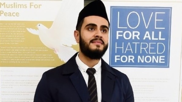 man with black beard in black muslim hat and suit and tie in front of pull up banner