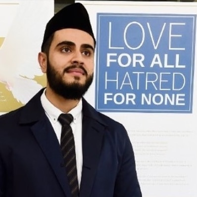 man with black beard in black muslim hat and suit and tie in front of pull up banner