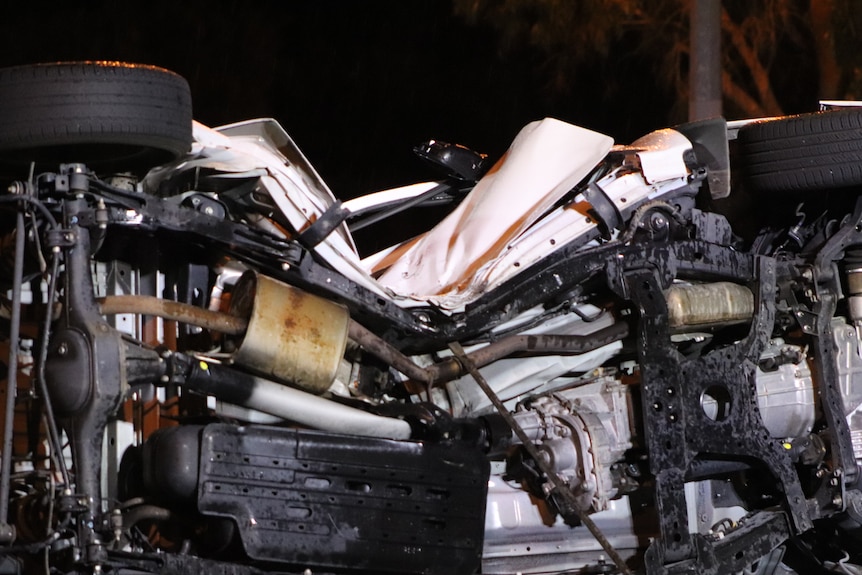 A close-up shot of the damaged driver's side and undercarriage of a white ute as it lies on its side after a crash.