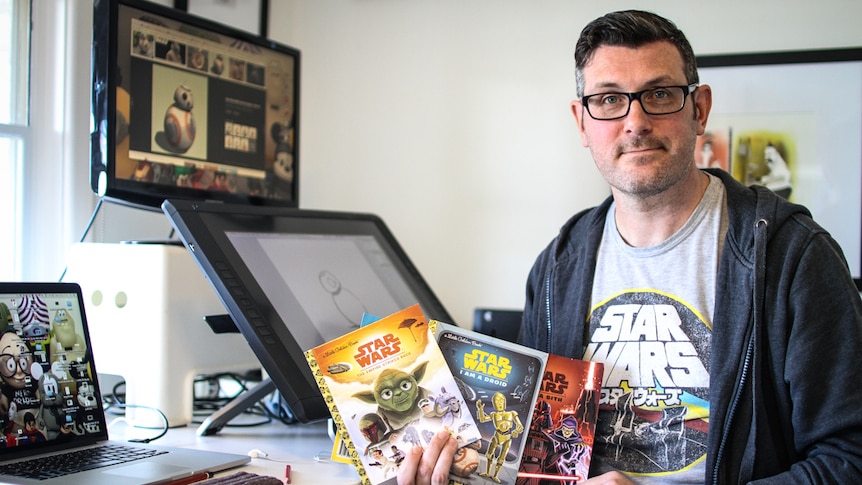 A man in a Star Wars t-shirt holding up three books.
