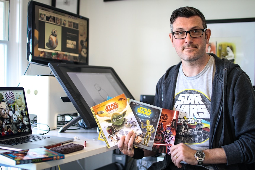 A man in a Star Wars t shirt holds up three books.