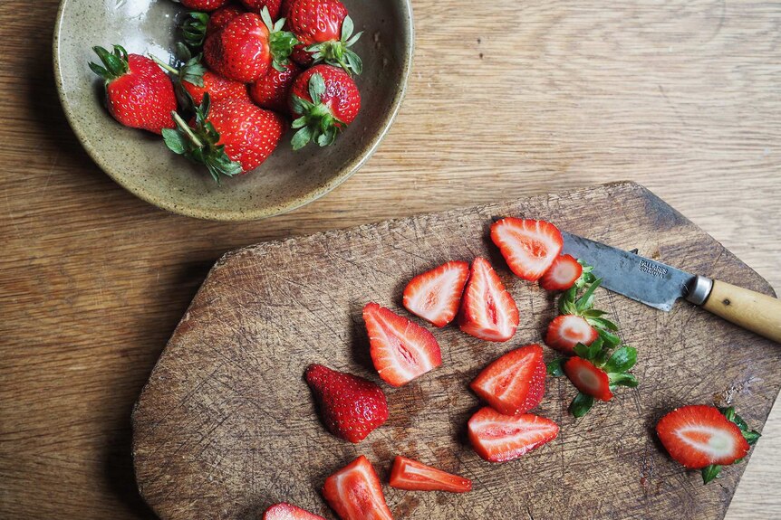 A bowl of fresh strawberries sits beside a chopping board where the berries are halved or quartered, for a shortcake filling.