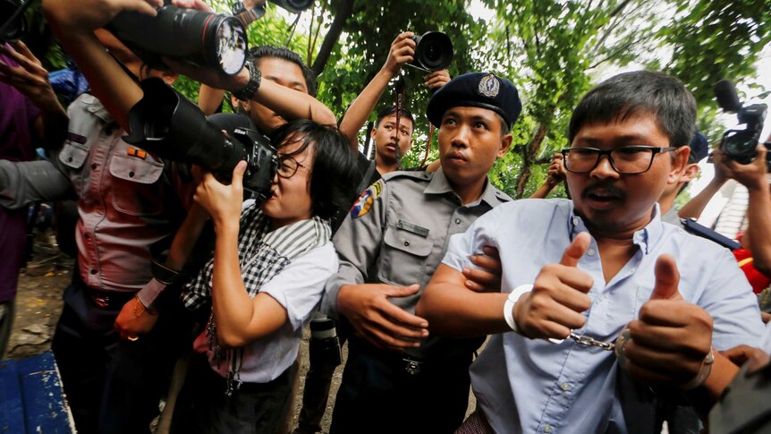 Detained Reuters journalist Wa Lone is escorted by police and is handcuffed. Nearby photographers point their cameras at him.