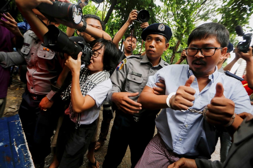 Detained Reuters journalist Wa Lone is escorted by police and is handcuffed. Nearby photographers point their cameras at him.