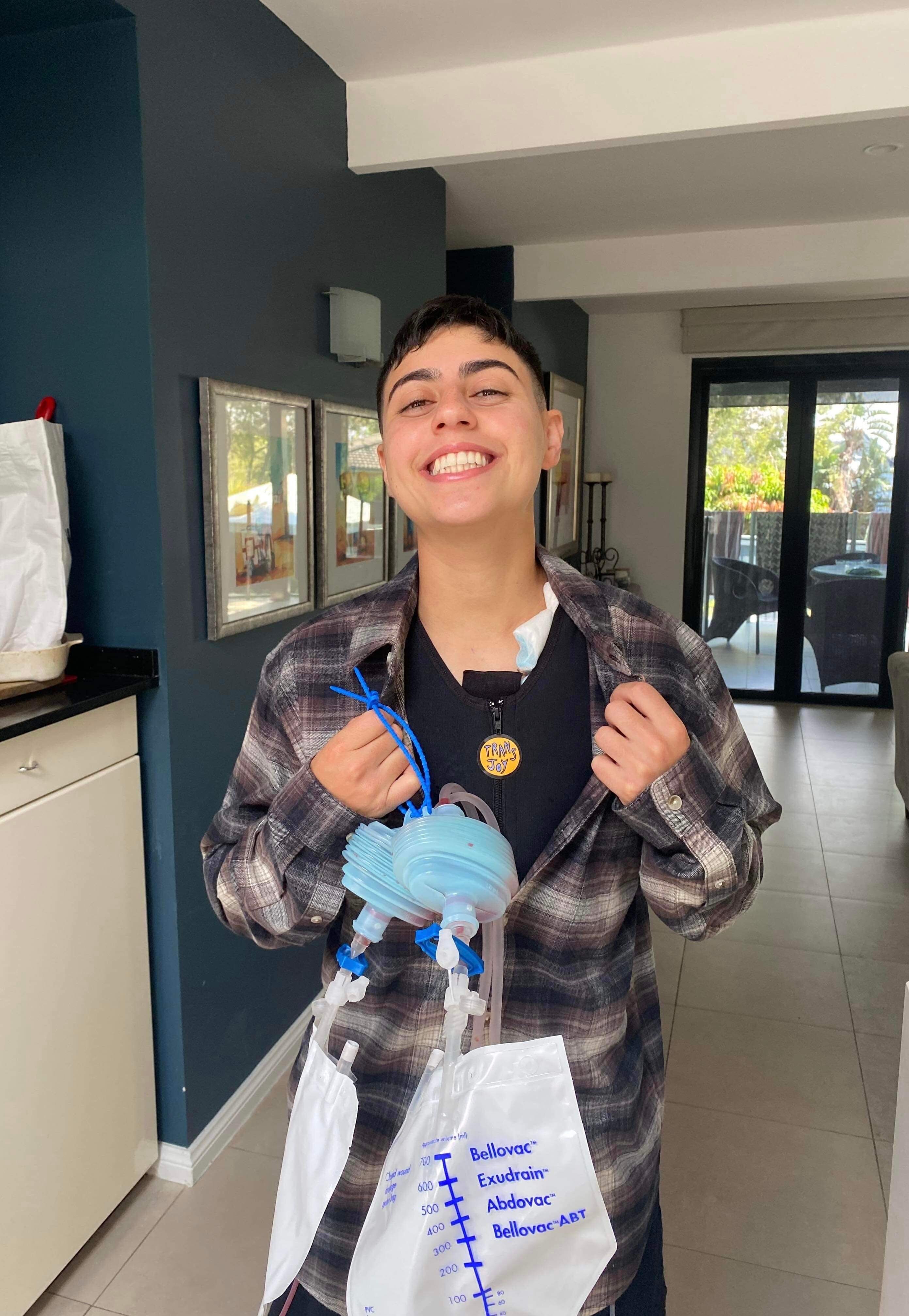 Speedy grins after surgery, holding their surgical drains and wearing a 'trans joy' badge.