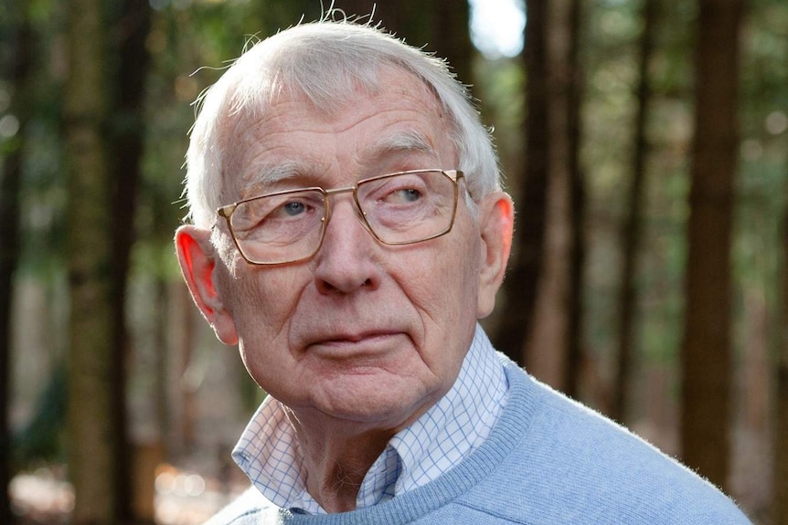 An old man in glasses and wearing a blue jumper stands in a forest.