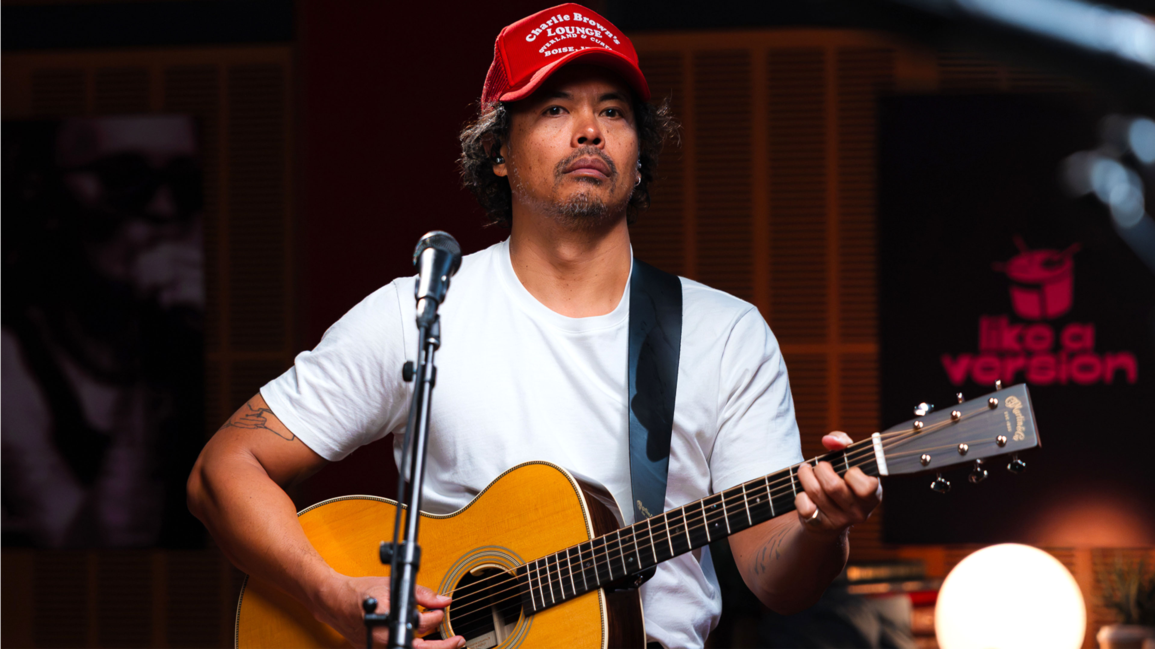 Dougy Mandagi wears a red ball cap and white t-shirt while holding an acoustic guitar