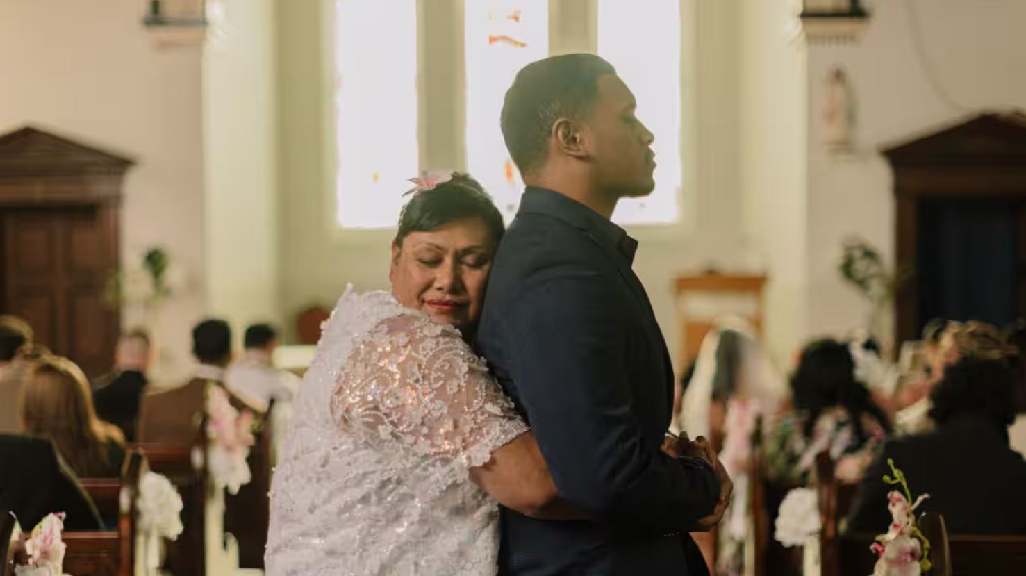 Samoan woman in a white dress hugs her son, wearing a suit, from behind. Stands in a church. 
