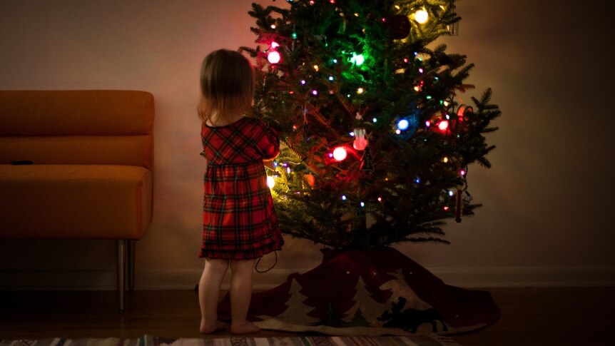 A small child decorates a green Christmas tree with rainbow lights glowing in a dim light.