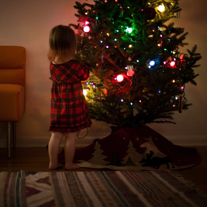 A small child decorates a green Christmas tree with rainbow lights glowing in a dim light.