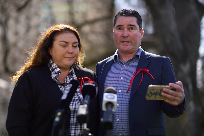 A woman and man wearing red ribbons stand in front of media microphones