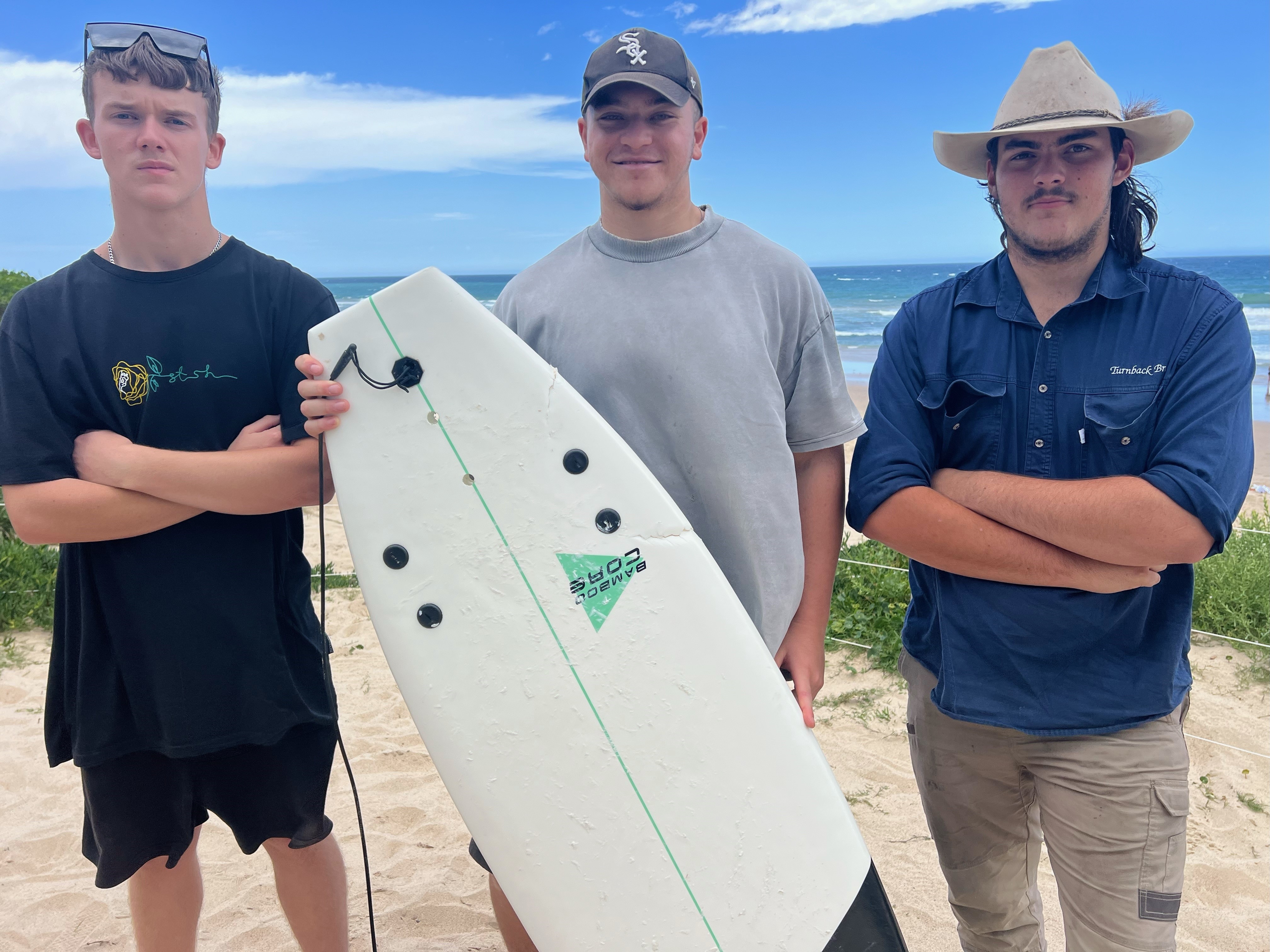 Julian McLennan standing with his friends and holding his surfboard, which has shark bite marks. 