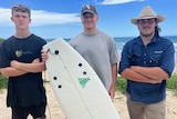Julian McLennan standing with his friends and holding his surfboard, which has shark bite marks. 