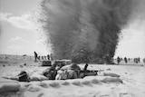 Black and white photo of a machine trench in the foreground and an explosion in the background