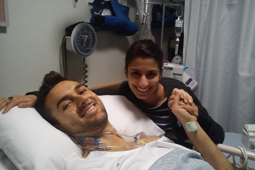 Ben Bravery lies in hospital bed after surgery with his wife next to him holding his hand