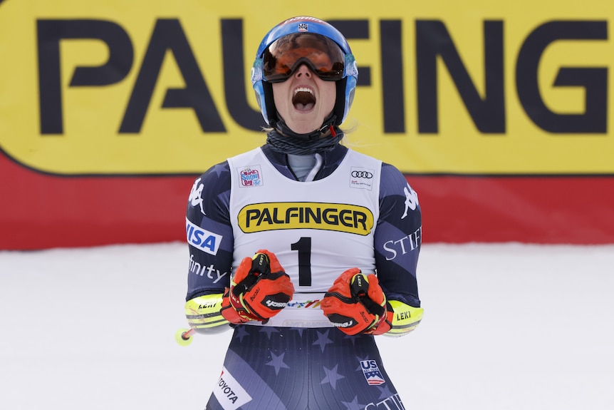 A skiier faces the camera, pumps her fists and roars with her helmet on after winning an event.
