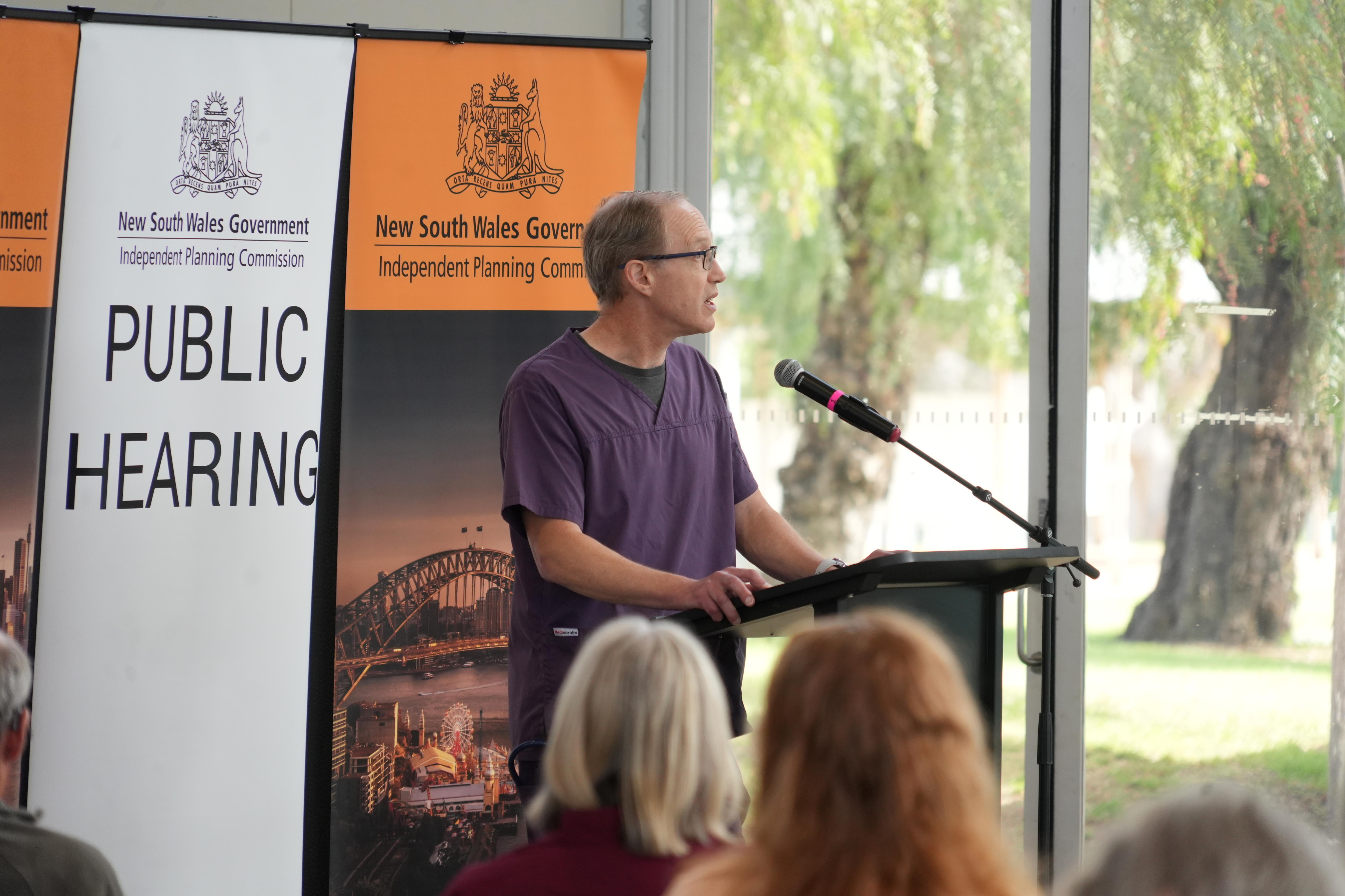 A man speaking at a lectern in front of a sign saying "public hearing".