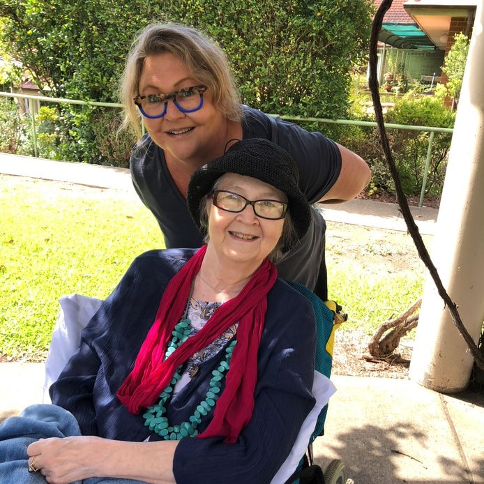 An older woman smiling in a wheelchair with her daughter behind her, leaning ovre her shoulder smiling