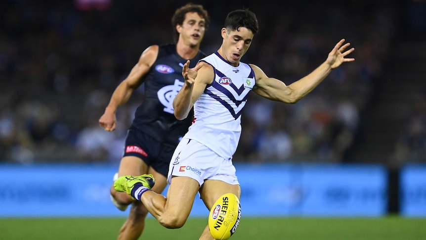 AFL player Adam Cerra in white away kit, kicking the ball with a Cats defender close behind him