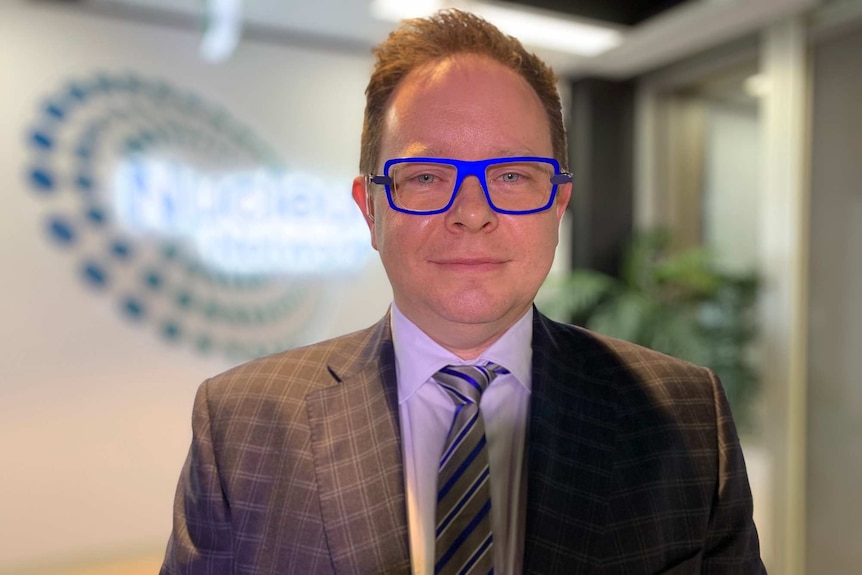 A man wearing blue-rimmed glasses and a suit stands in an office foyer.