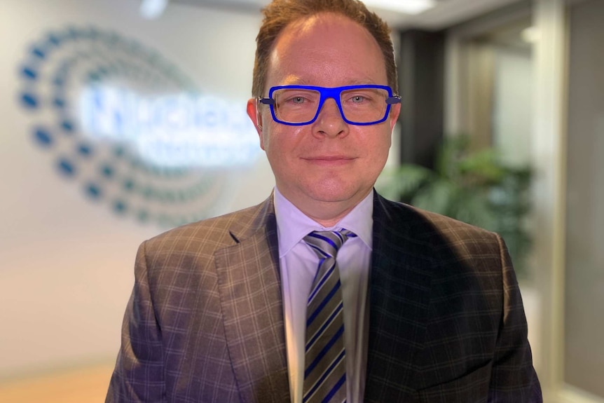 A man wearing blue-rimmed glasses and a suit stands in an office foyer.