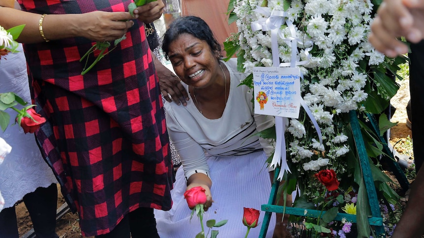 A woman in white weeps on her knees surrounded by roses and white flowers.