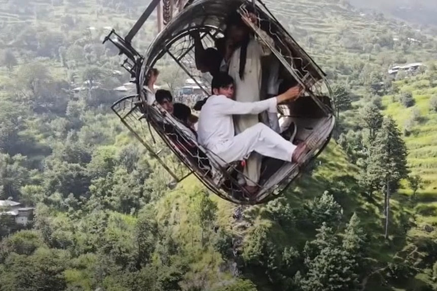 Multiple teenage boys crouch inside a cable car dangling at an angle above a forest. 