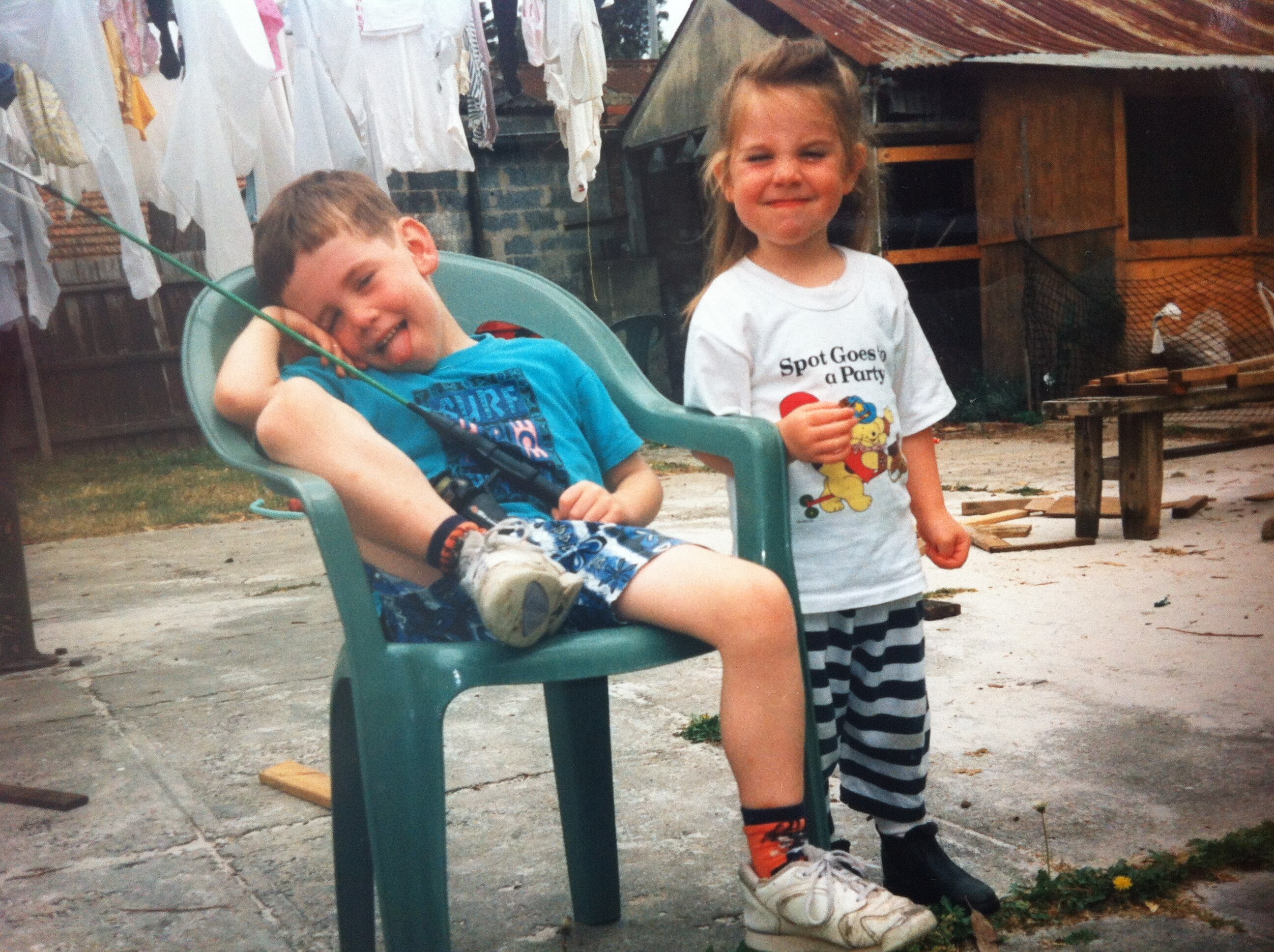 Angie, left, stands and pulls a face as she stands next to her brother, left, who sits on a chair in a concrete backyard.