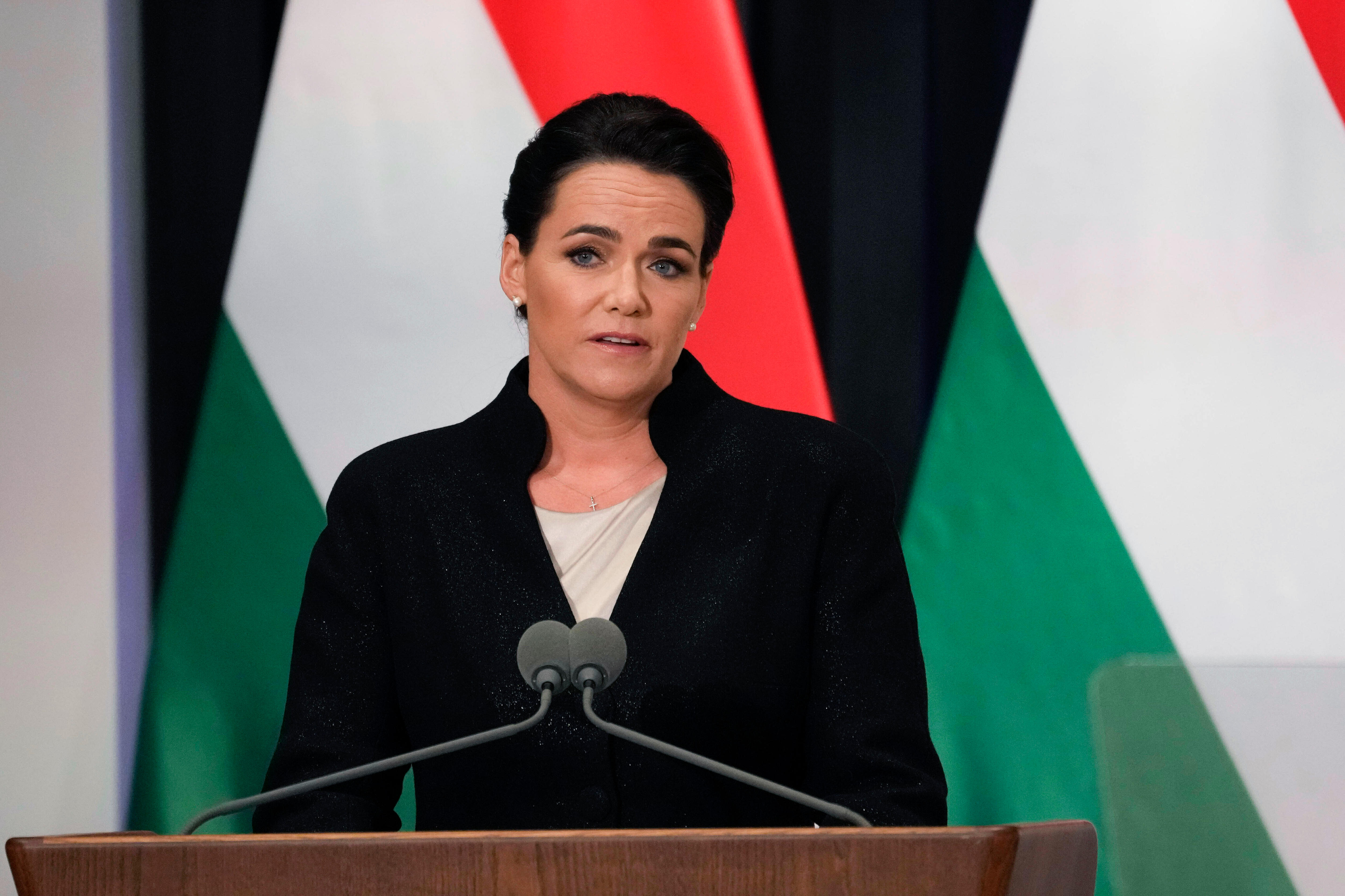 A middle-aged woman with dark hair speaks at a podium with Hungarian flags behind her.