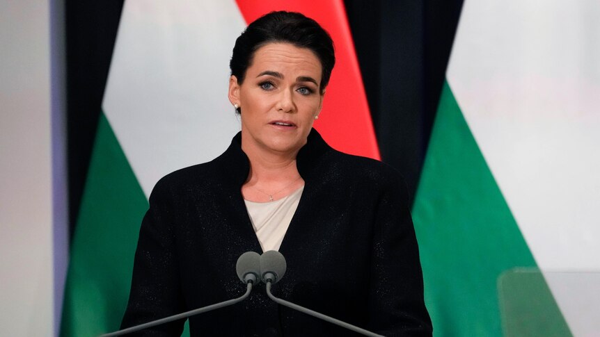A middle-aged woman with dark hair speaks at a podium with Hungarian flags behind her.
