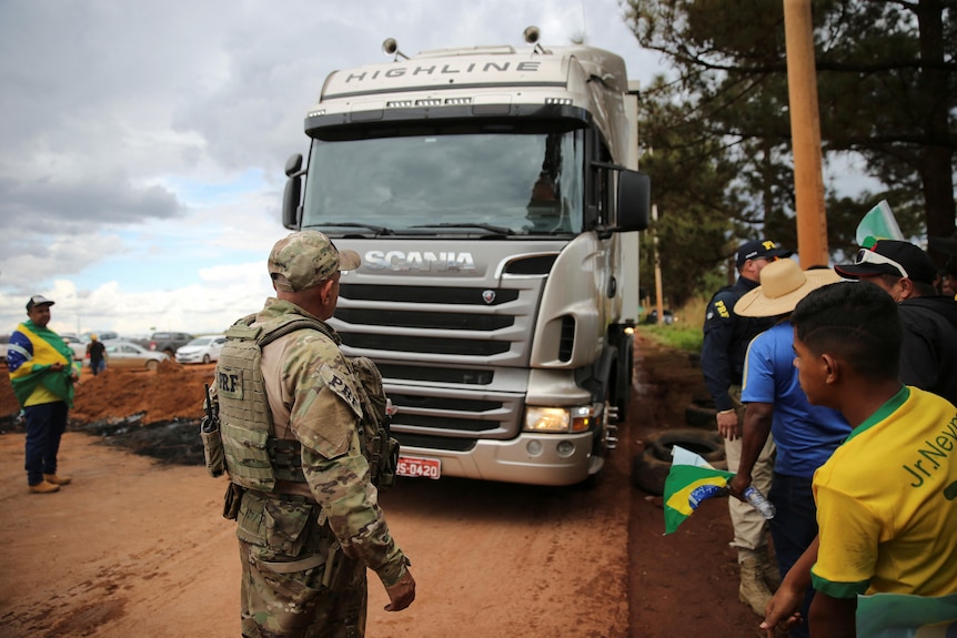 A man dressed in military fatigues stands in front of a truck with people dressed in yellow shirts watching nearby.