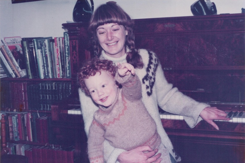 an old image of a woman with pigtail plaits holding a young boy in a jumper, a piano in the background