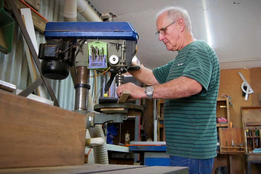 A man in his 60s uses a large drill in a backyard work shed