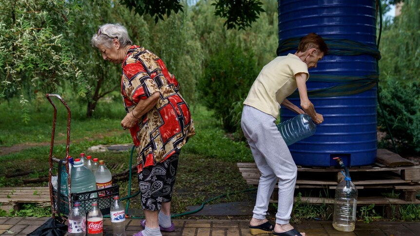 Two older woman stand near a water tank, filling bottles and containers 