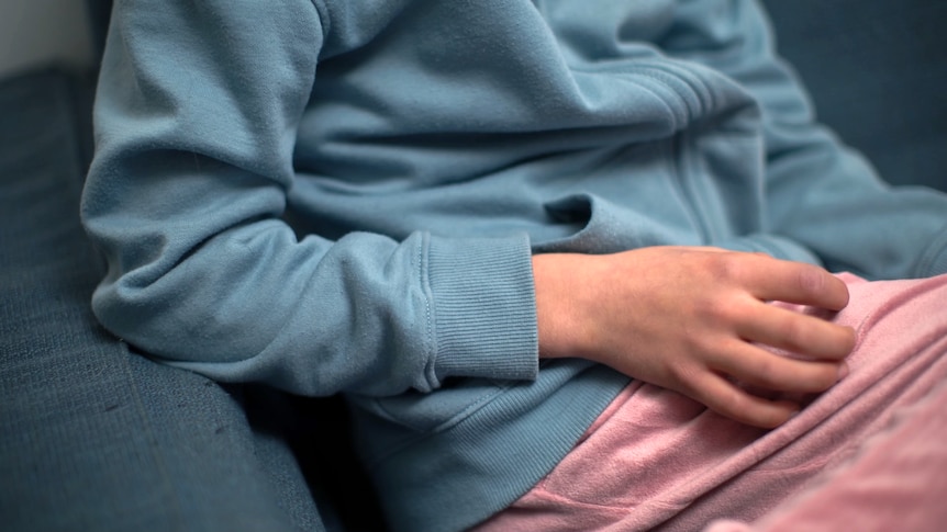 An unidentifiable child wearing a blue jumper and pink pants sits on a couch. They are resting a hand on their lap.