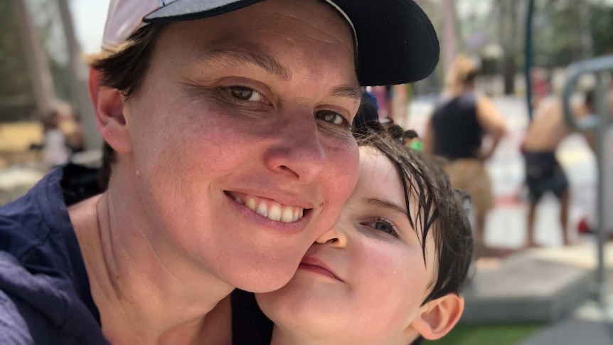 A woman wearing a cap and holding her young son smiles for the camera. Her son wears a white swim shirt.