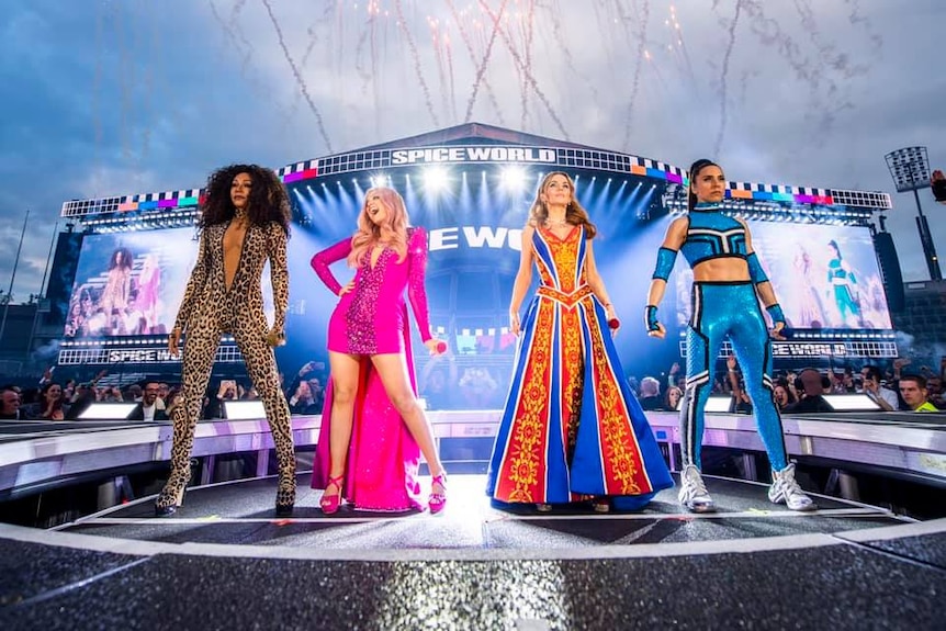 Scary, Baby, Ginger and Sporty Spice standing on a stage surround by fans, fireworks in the background.