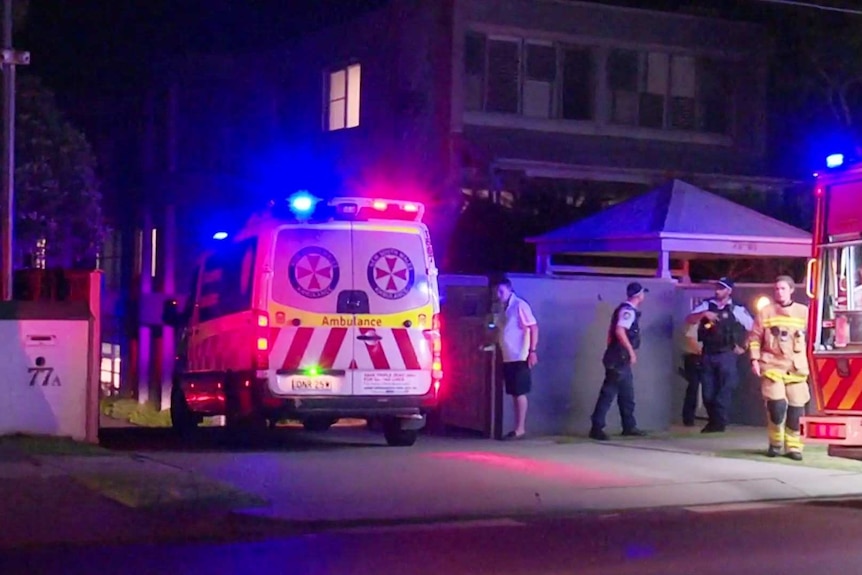 Une ambulance pénètre dans une allée la nuit tandis que les pompiers et la police se tiennent à proximité.