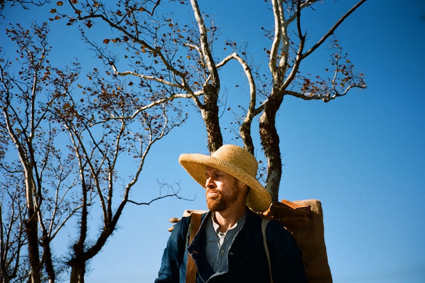 Colour still of Willem Dafoe standing outside in front of trees with autumn leaves and blue sky in 2018 film At Eternity's Gate.