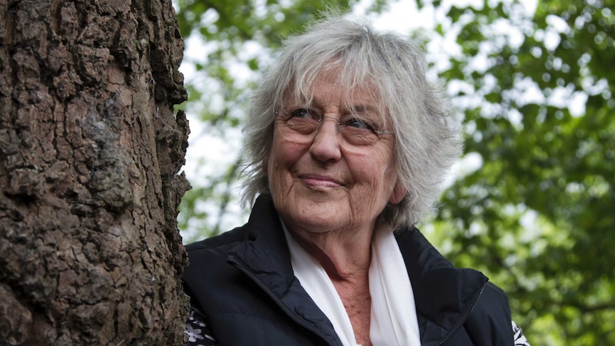 Legendary Australian feminist writer Germaine Greer, standing next to a tree, smiles as she looks up into the sky.