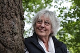 Legendary Australian feminist writer Germaine Greer, standing next to a tree, smiles as she looks up into the sky.