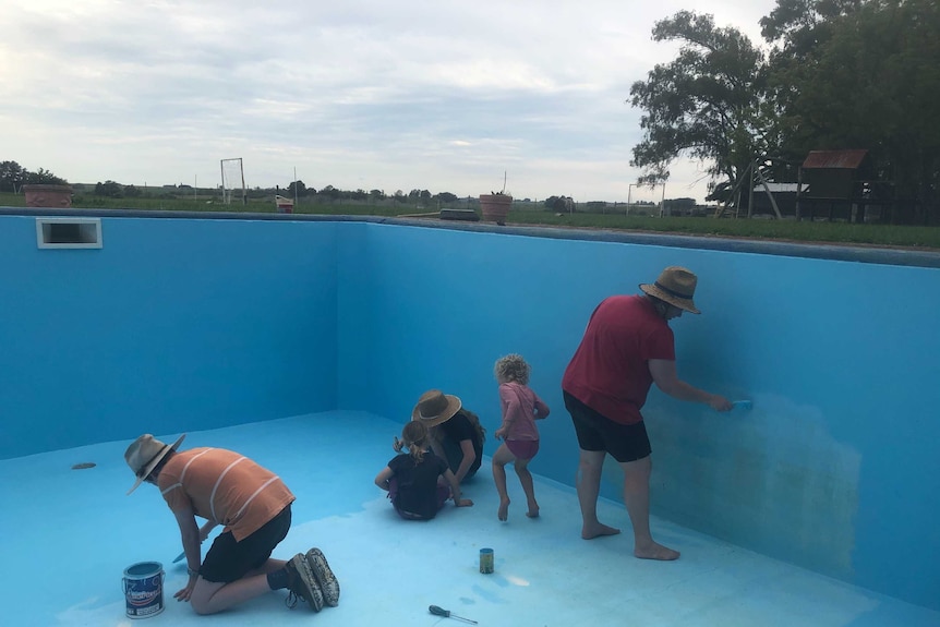 A mix of adults and children, most wearing hats, in an empty pool painting the sides blue.