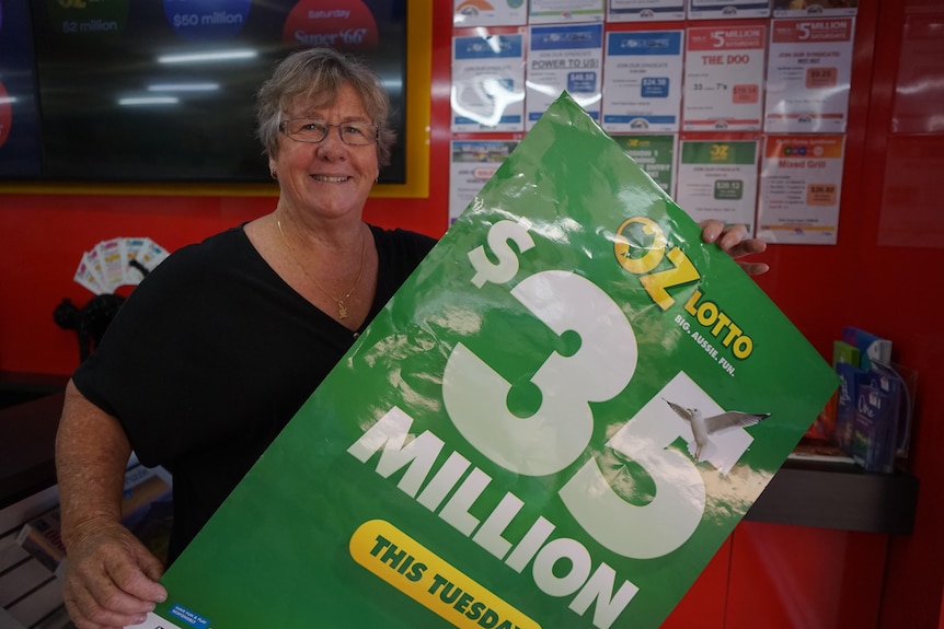 Newsagent Kerry Magree stands holding a large green poster that says: 'OzLotto $35 million' in white writing.