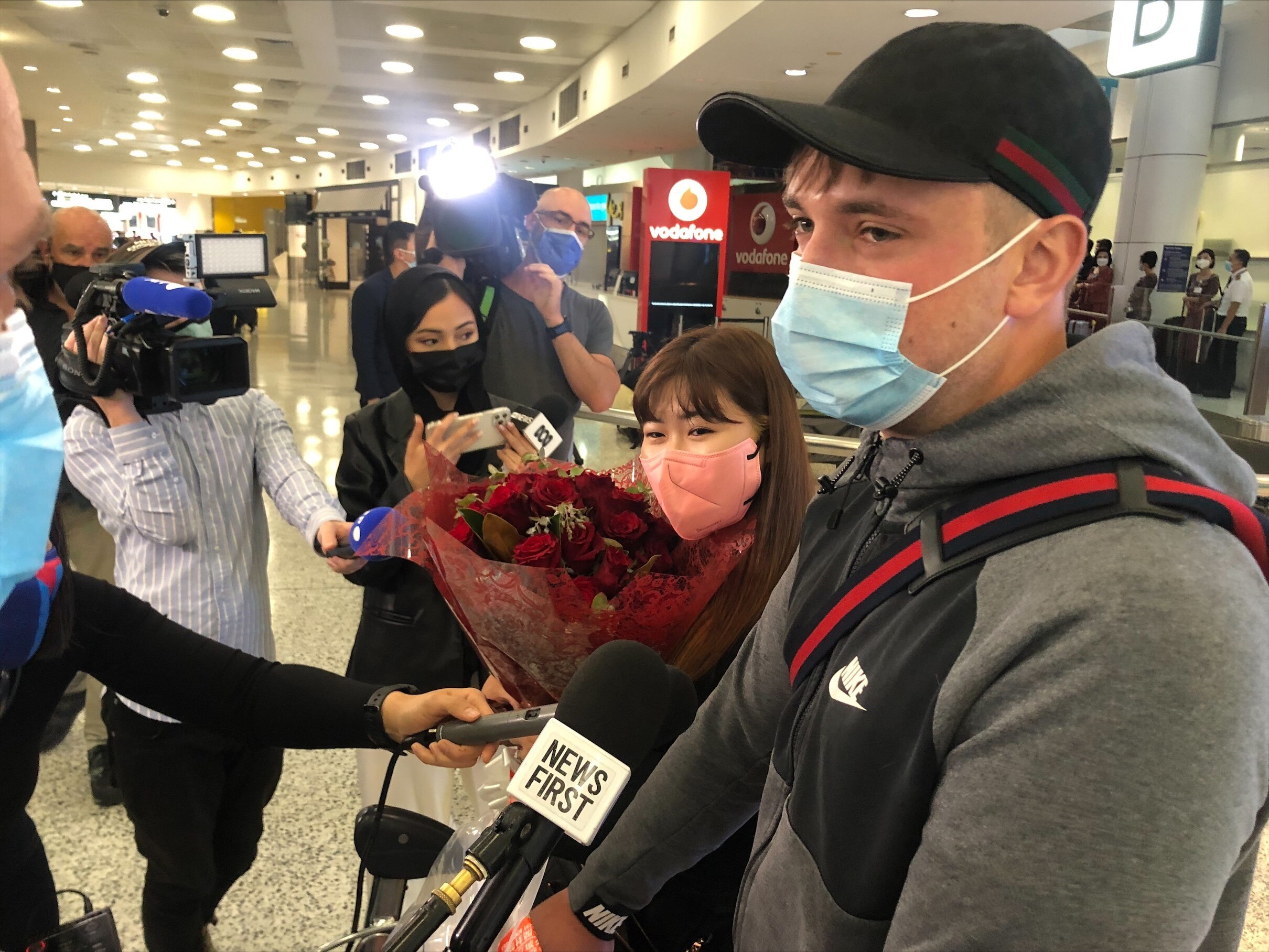 A man and a woman holding a bunch of roses are surrounded by reporters holding out microphones. 