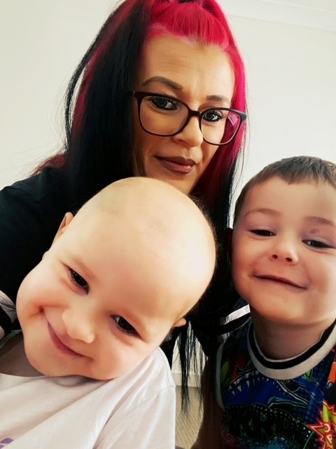 Una mujer pelirroja con gafas posa con dos niños pequeños, incluido uno sin pelo tras un tratamiento contra el cáncer.