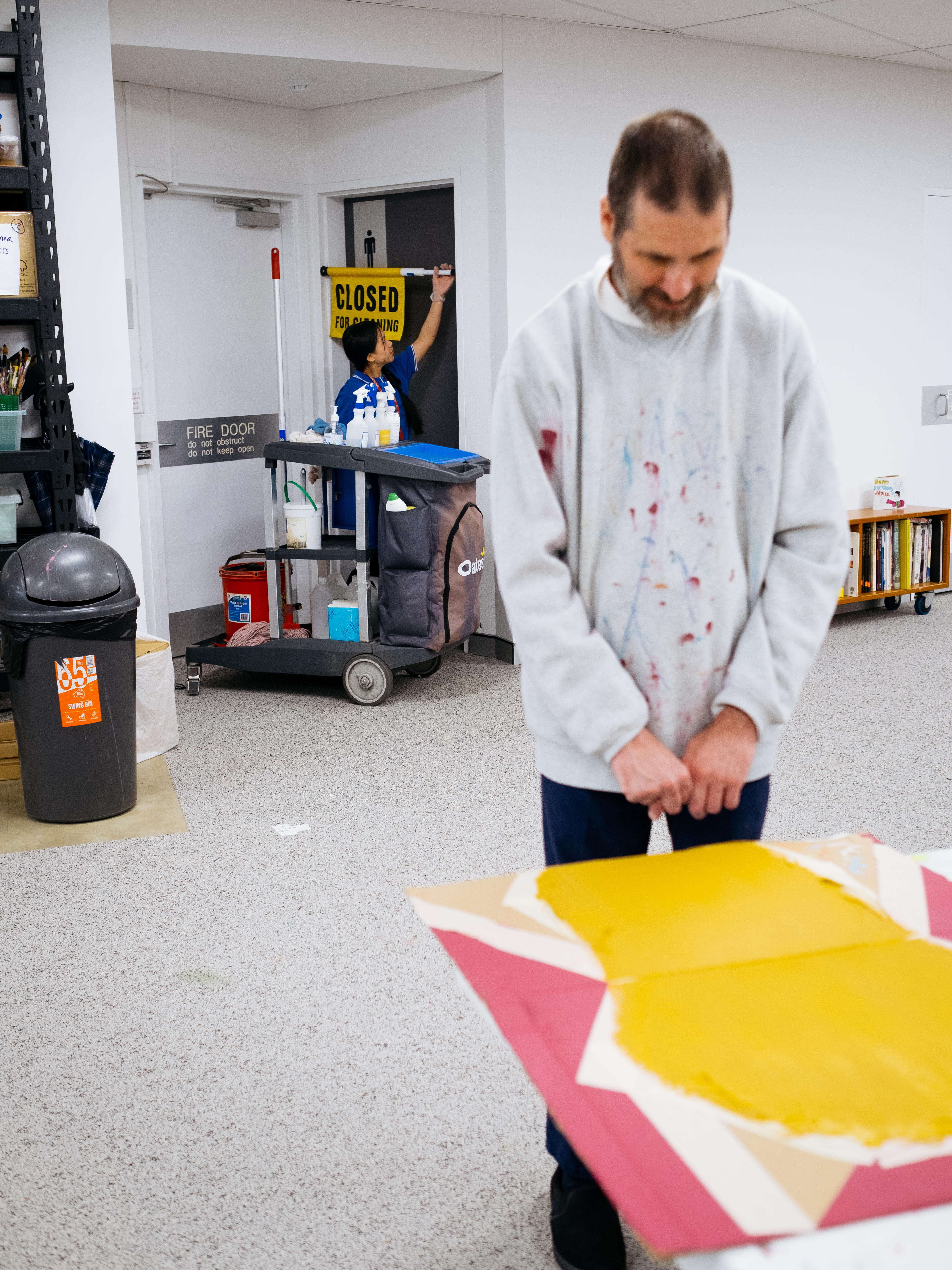 Artist Thom painting at a table in a grey jumper, with cleaner Subita and her trolley leaving the bathrooms in the background