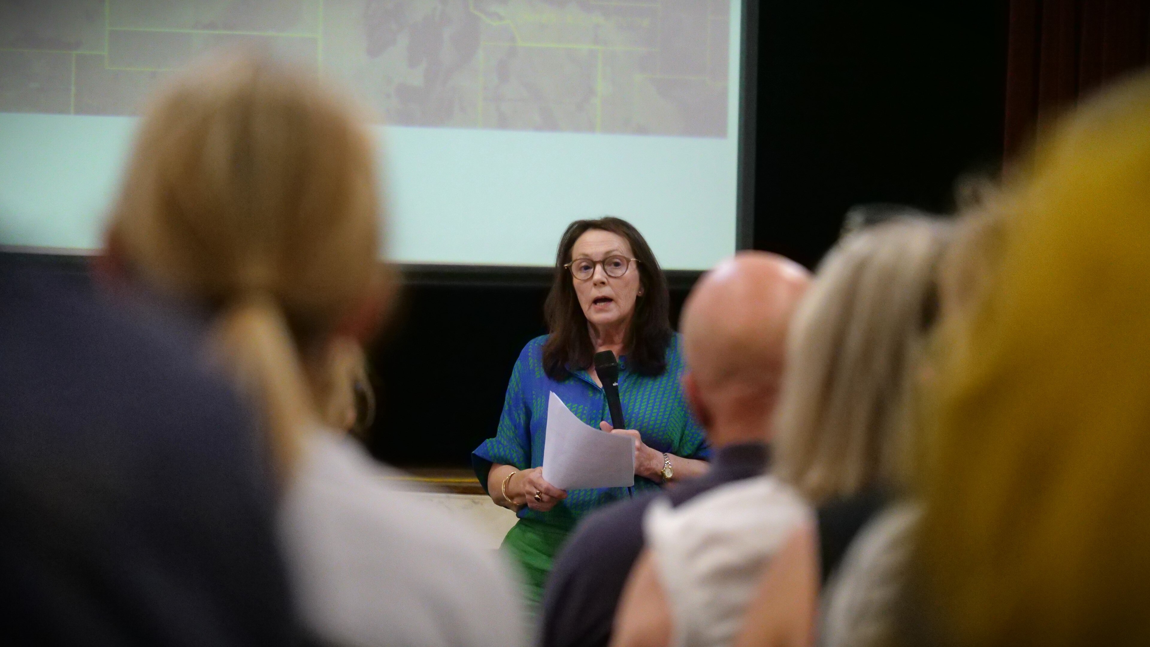 A woman in a blue shirt speaks to a crowd of people holding a microphone and paper. 