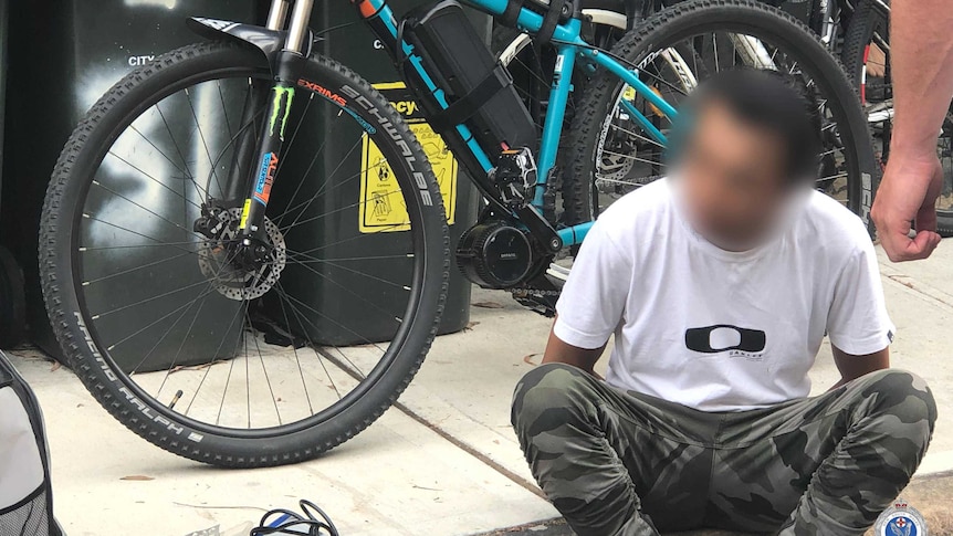 A man with a blurred face sits on the ground in front of a bicycle.