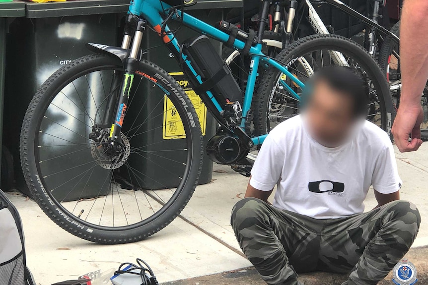 A man with a blurred face sits on the ground in front of a bicycle.