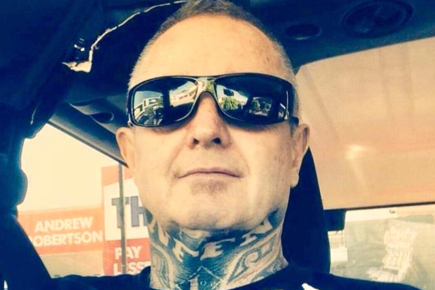 A bald man with a tattooed neck sits in a car, wearing dark sunglasses.